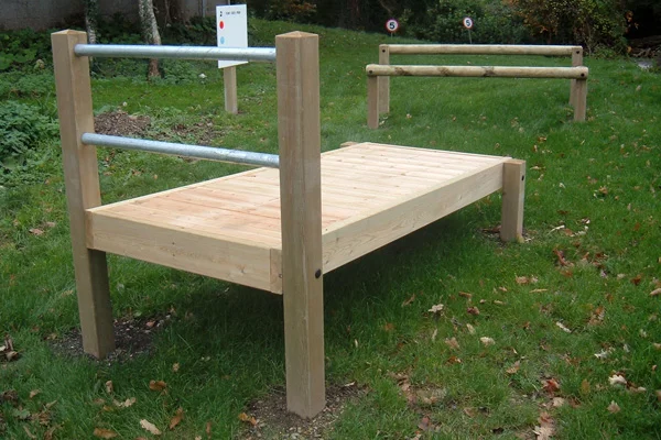 A wooden Abdominal Exerciser outdoor fitness equipment with a flat timber platform and galvanised steel bar, installed on a grass park or school field, with a second unit visible in the background.