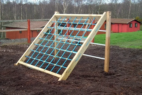 An A-frame timber assault net with a blue rope net and a ladder, set in a loose-fill earth and bark safety surface in a rural outdoor playground.