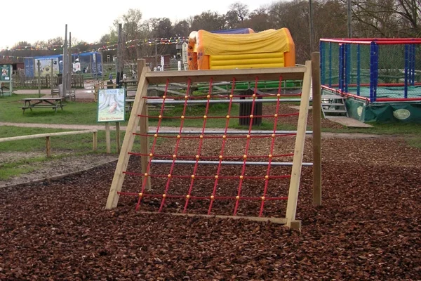 A red rope Assault Net climbing frame installed on a wood chip playground surface at a visitor attraction, with colourful play equipment visible in the background.