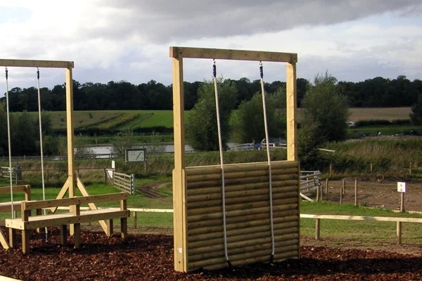 A timber log assault wall with two climbing ropes, part of an extensive outdoor activity course with other trim trail equipment, on a bark mulch safety surface in a rural setting.