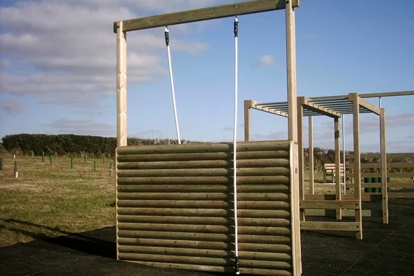 A timber log assault wall with two climbing ropes, part of an outdoor trim trail at an activity centre, set on a black rubber matting surface in a grassy field.