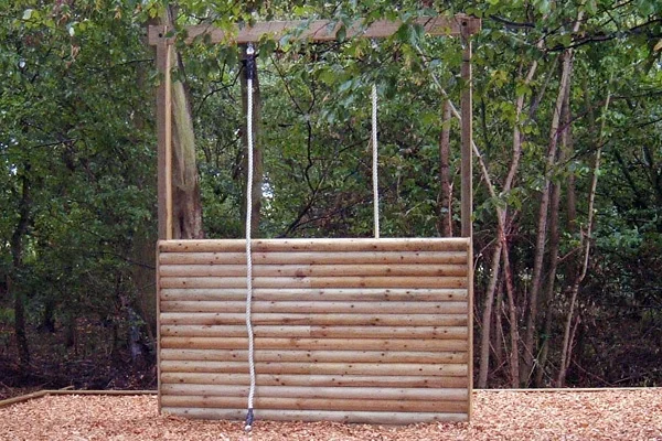 A timber log assault wall with a single climbing rope, set in a woodland area on a bark mulch safety surface, as part of a trim trail.