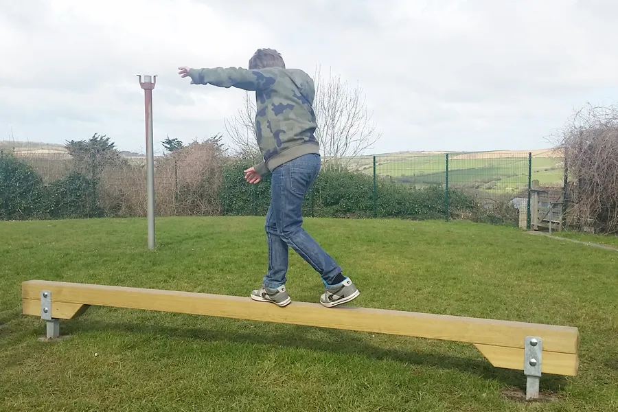 A boy in a camouflage hoodie balancing on a wooden Balance Beam with his arms outstretched, on a grass outdoor play area with rolling hills in the background.