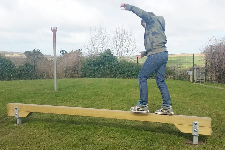 A boy in a camouflage hoodie balancing with arms spread wide on a wooden Balance Beam on a grass school playground.