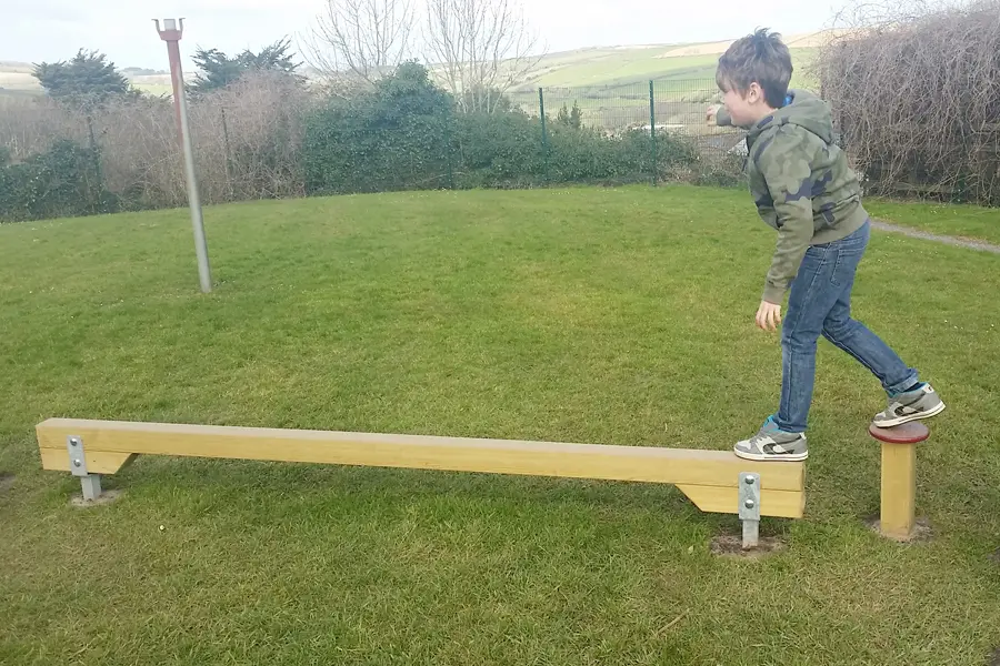 A boy stepping from a wooden Balance Beam onto a small red-capped balance post at the end of the beam, on a grass school playground.
