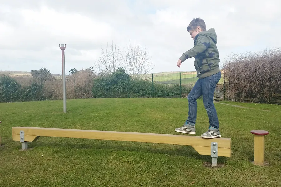A boy in a camouflage hoodie looking down as he carefully walks along a wooden Balance Beam on a school field, with a small red-capped balance post visible at the end.