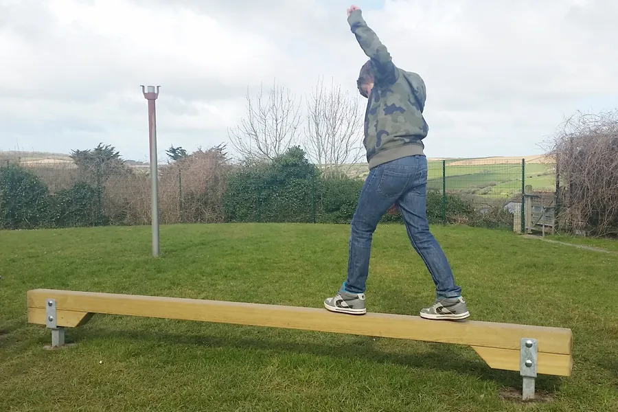 A child in a camouflage hoodie walking along a wooden Balance Beam with one arm raised, on a grass outdoor trim trail with countryside views.