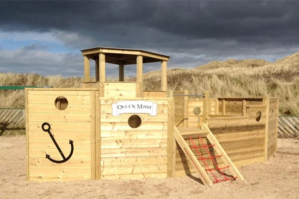 The 'Queen Mary' Beachside Boat play ship on a sandy beach, with a climbing net and a backdrop of dunes and a dramatic sky.