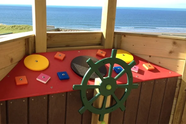 A view from the helm of the Beachside Boat, showing the ship's wheel, a play panel, and a stunning ocean backdrop.