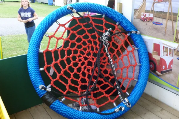 A young girl sitting inside a large bird nest swing with a blue rim and red rope net, which is on display in a van at a trade show.