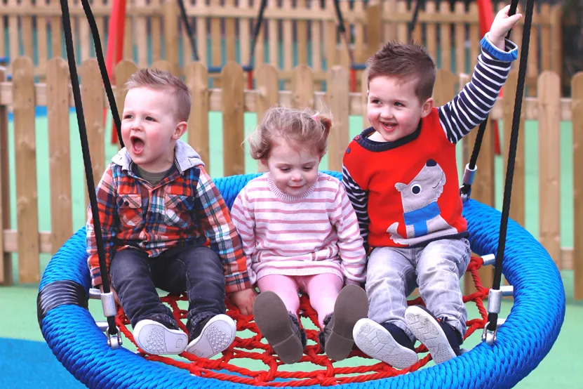 Three toddlers sitting together in a large bird nest swing with a blue rim and red rope net, in a playground with a wooden fence and a colourful rubber surface.