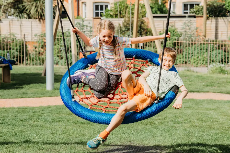 A boy and a girl laughing together on a large, round bird nest swing with a blue rim and red rope net, in a sunny, grassy outdoor playground.