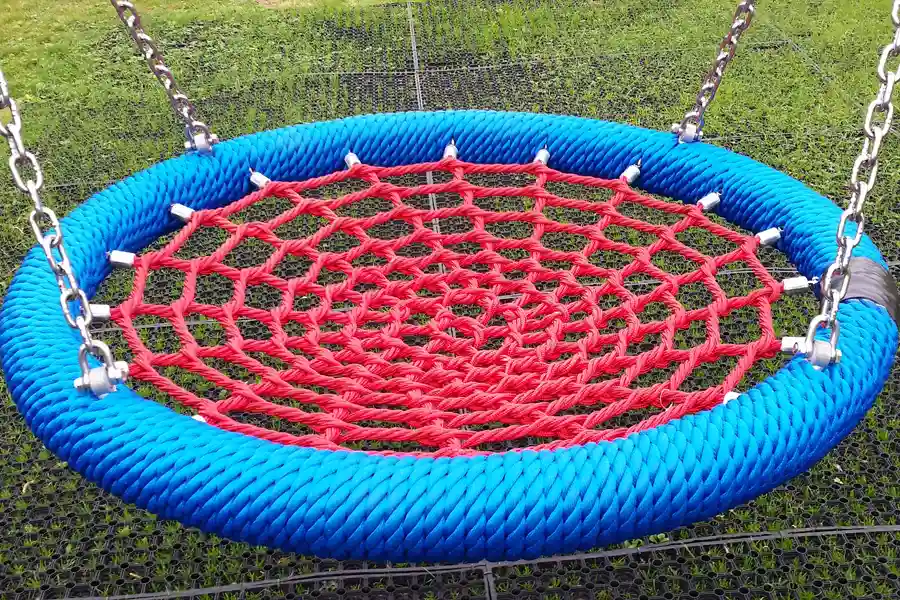 A wide-angle product shot of a bird nest swing seat with a blue rope rim and red rope net, suspended by steel chains over a black rubber grass mat.