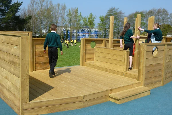 Schoolchildren in uniform playing on the HMS St Meriadoc, a timber boat stage with a decked platform, portholes, and posts, on a blue rubber safety surface in a school playground.