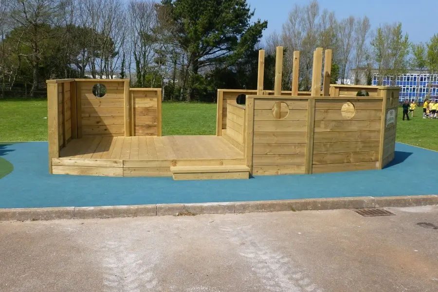 A wide view of a large timber boat stage play structure with a decked stage, portholes, and mast-like posts, set on a blue rubber safety surface in a school playground.