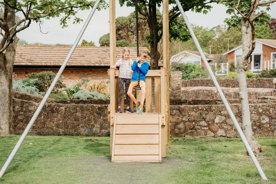 A girl and a boy standing on the wooden launch platform of a Cableway zip wire, with the boy gripping the zip wire handle, in a garden playground setting.