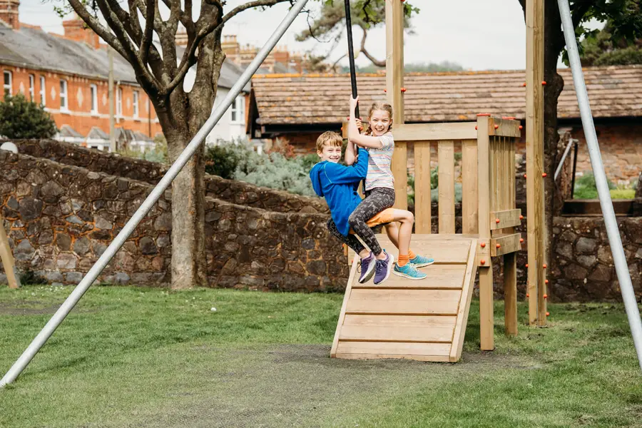 Two smiling children a boy in a blue hoodie and a girl in a striped top swinging together on a Cableway zip wire at an outdoor playground, with a stone wall and trees in the background.