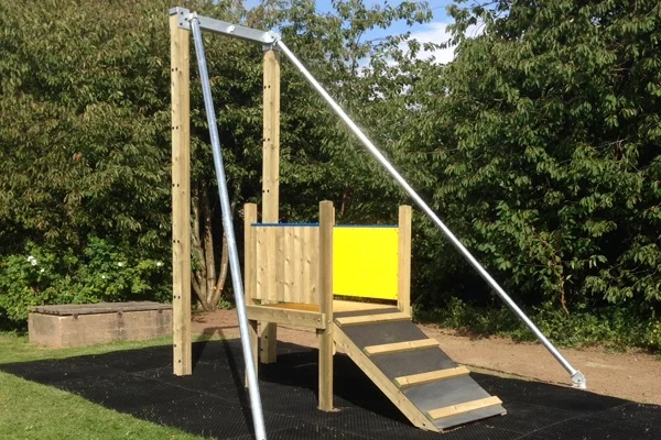 A close-up side view of a Cableway zip wire showing the wooden launch platform with yellow safety panel, galvanised steel A-frame, and rubber matting in a sunny garden playground.