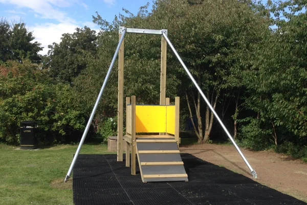 A Cableway zip wire with a wooden launch platform, yellow safety panel, and galvanised steel frame, installed on rubber matting in a sunny school playground surrounded by trees.