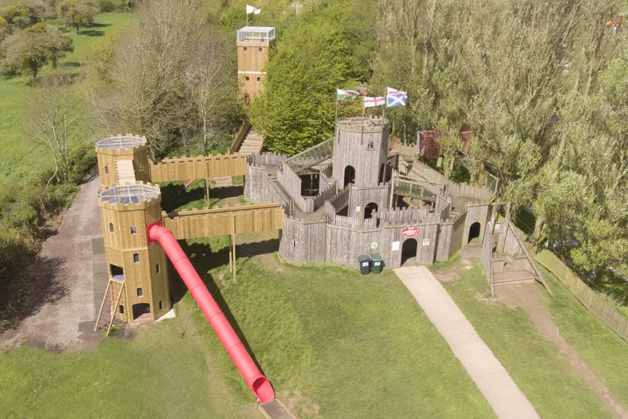 An aerial view of the Cattle Country Fort, a large timber play castle with multiple towers, a long red tube slide, elevated walkways, and flags, set in a grassy outdoor playground surrounded by trees.