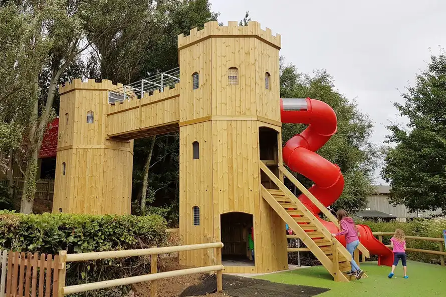 Two children playing on the Cattle Country Fort, a timber play castle with a red spiral tube slide, a wooden staircase, and a walkway connecting two towers, in a wooded outdoor playground.