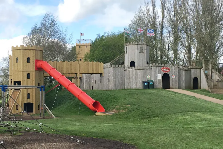 The Cattle Country Fort, a timber play castle with a large red tube slide, battlements, and flags, with a sign for The Real Berkeley Castle, in a grassy outdoor playground setting.