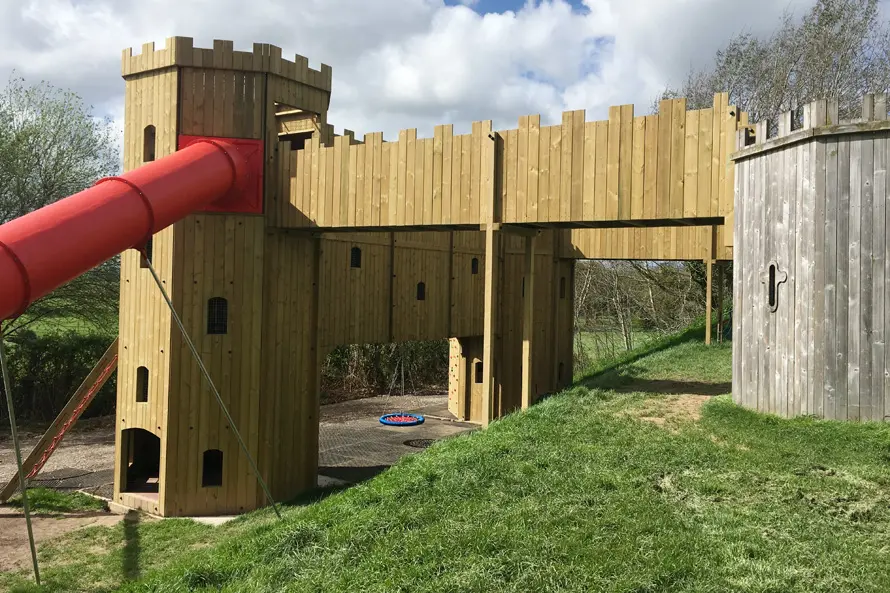 A view of the Cattle Country Fort showing the red tube slide, a timber walkway, and a nest swing, with the fort set on a grassy mound in an outdoor playground.