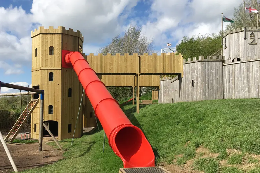 The red tube slide of the Cattle Country Fort, emerging from a timber tower with battlements, with a wooden walkway and another tower with flags visible in the background, in a grassy outdoor playground.