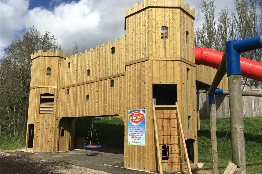 A close-up of the Cattle Country Fort, a timber play castle showing a rock climbing wall, a nest swing, a rope ladder, and the entrance to the red tube slide, with a sign for The Real Berkeley Castle.