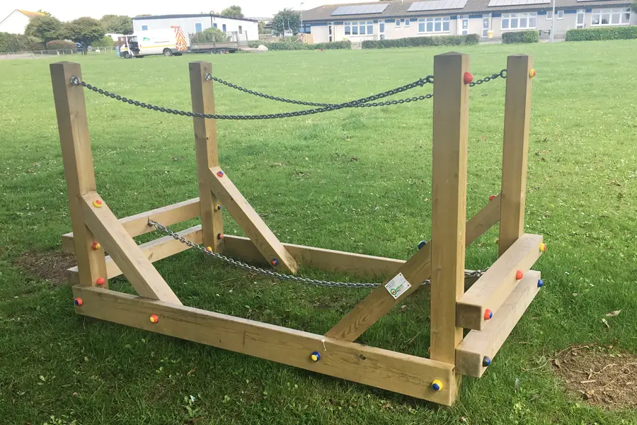 A three-quarter view of a wooden Chain Bridge outdoor play equipment with chain handrails on a grass school playground, with school buildings visible in the background.