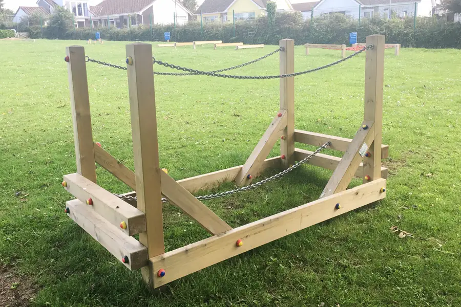 An angled view of a wooden Chain Bridge trim trail equipment with chain handrails and coloured bolt caps on a school field, with other trim trail equipment visible in the background.