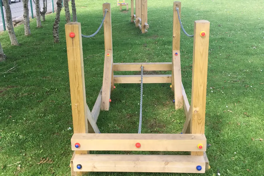 A front view of a wooden Chain Bridge outdoor play equipment with chain handrails and coloured bolt caps, installed on a grass school playground.