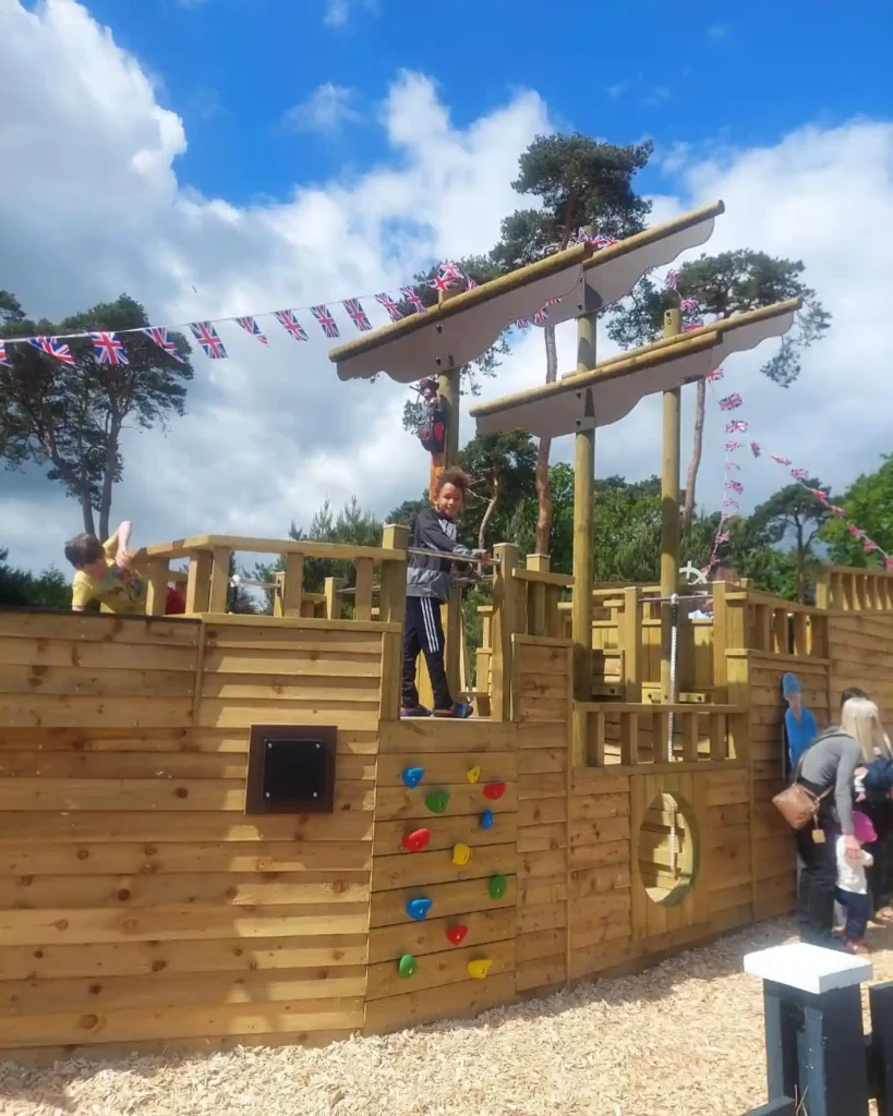 Children playing on The Coverack Queen, a large wooden outdoor play ship with a climbing frame, at a playground under a blue sky.