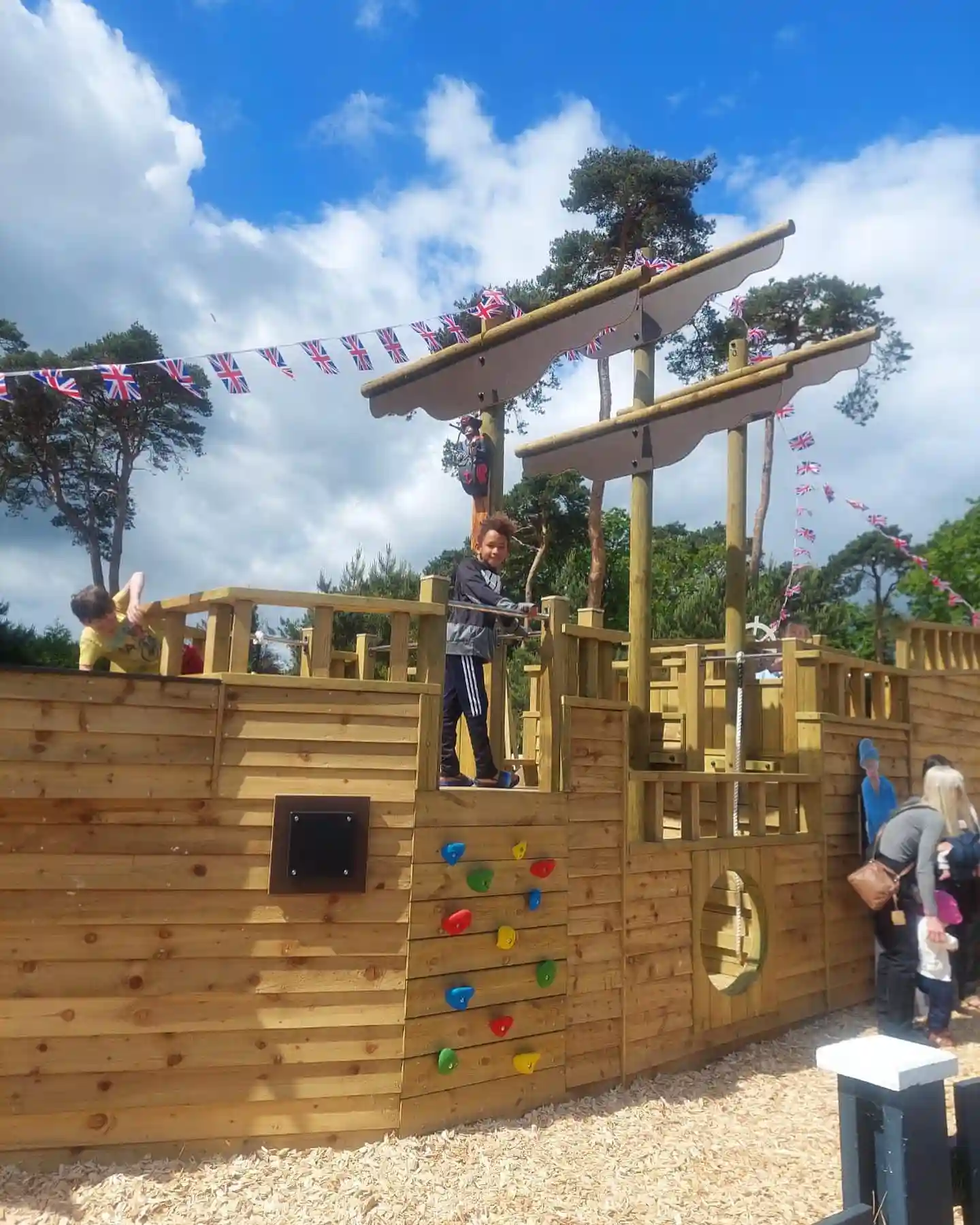 Children playing on The Coverack Queen, a large wooden outdoor play ship with a climbing frame, at a playground under a blue sky.