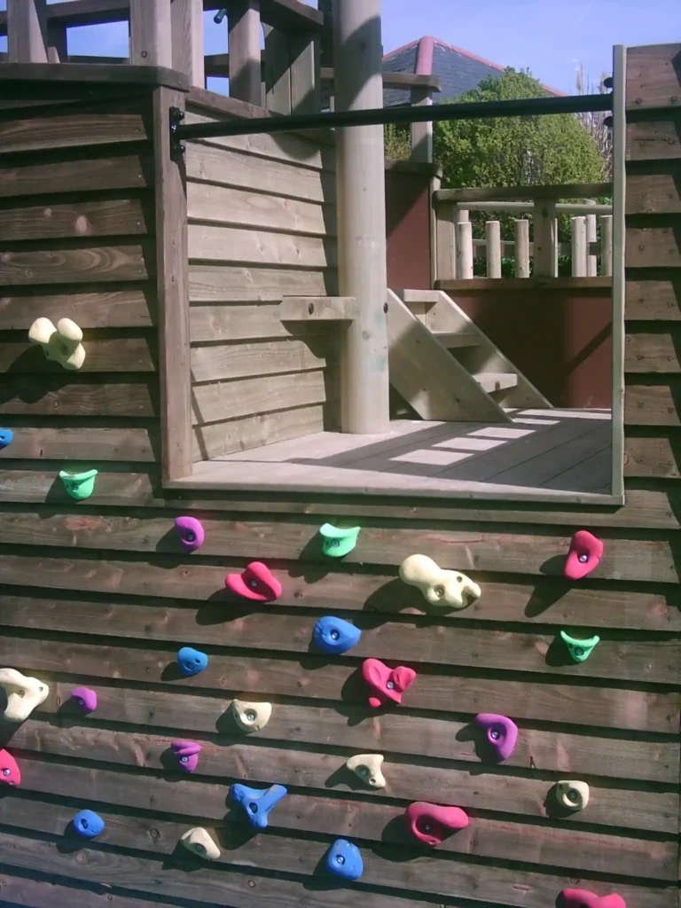 A colourful climbing wall with various holds on the side of The Coverack Queen outdoor play ship, leading to the deck.