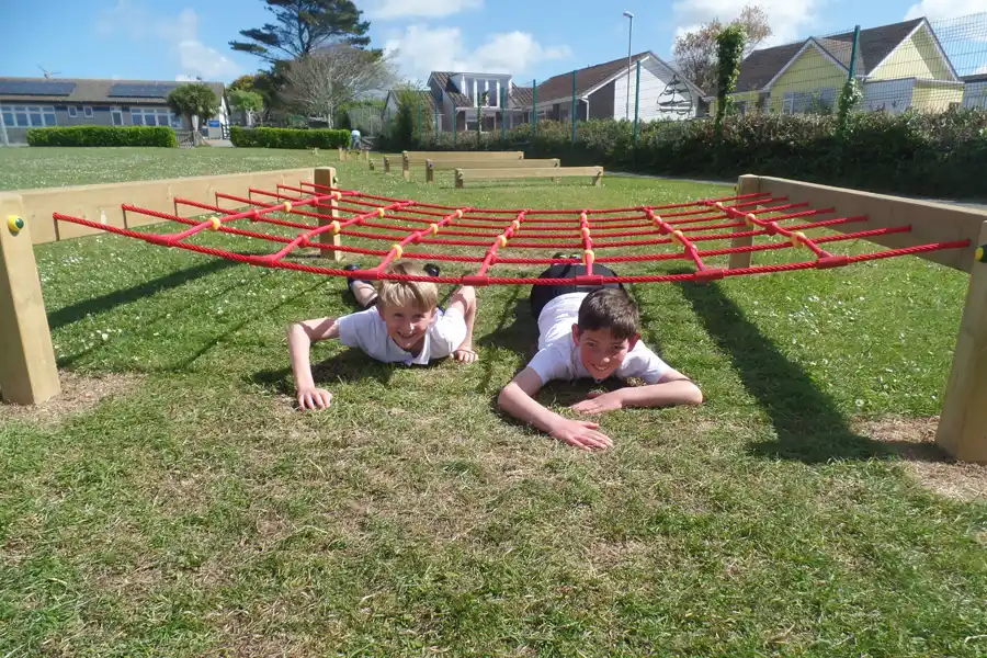 Two boys in school uniform crawling under a red rope Crawl Net on a sunny school playground, smiling at the camera.