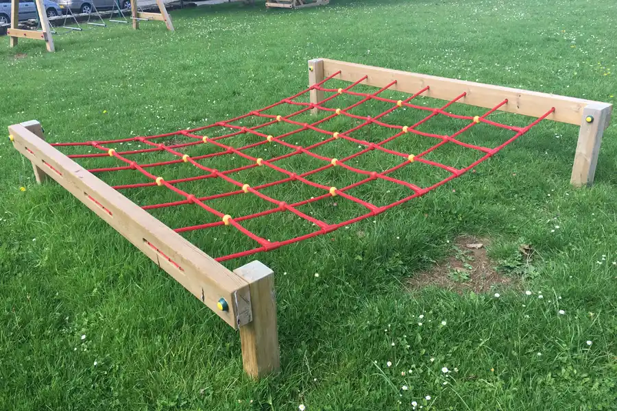 A red rope Crawl Net outdoor play equipment with yellow connectors, suspended between a wooden frame on a grassy school field.