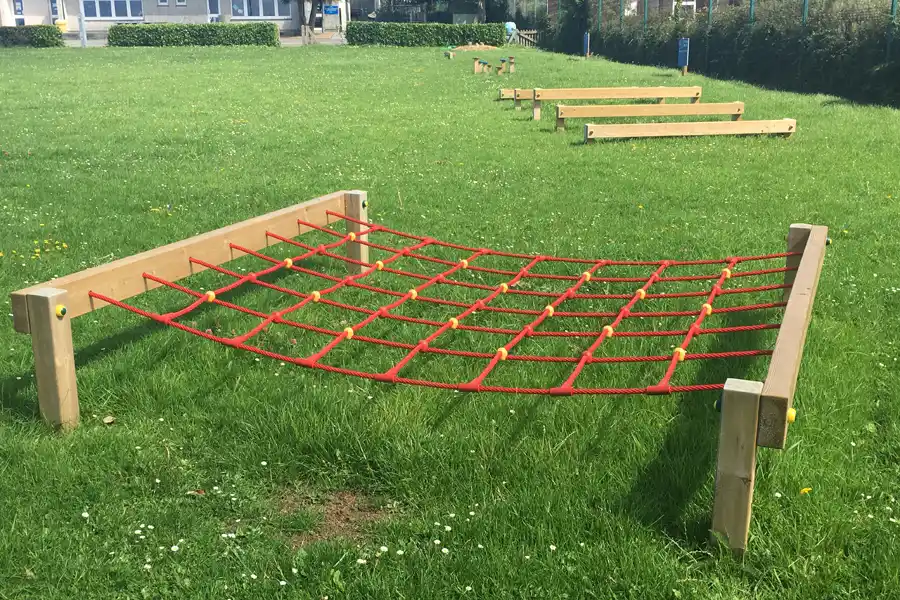 A red rope Crawl Net installed on a grass playground as part of an outdoor trim trail course, with other trim trail equipment visible behind.