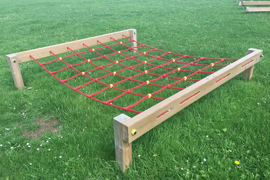 A side view of a red rope Crawl Net trim trail equipment on a school field, with more trim trail equipment visible in the background.