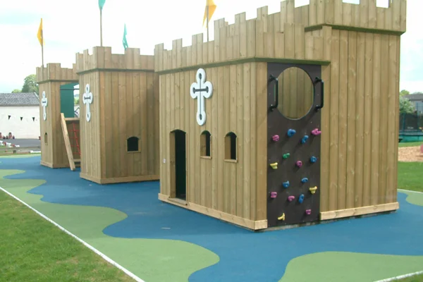 The Dalscone Fort timber play castle from another angle, showing the rock climbing wall, a circular entrance, a rope ladder, and castle battlements, on a blue and green rubber safety surface.