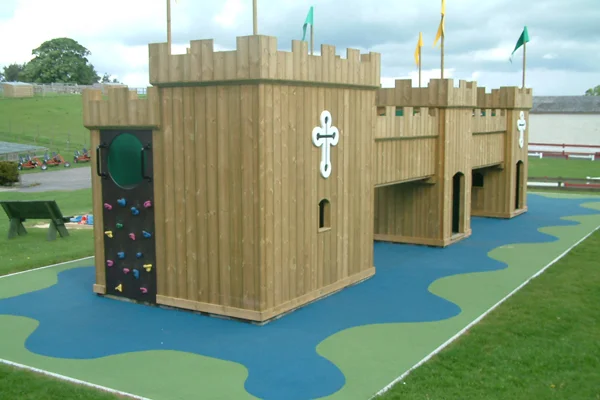 A timber play castle, the Dalscone Fort, with a rock climbing wall featuring colourful holds, a circular porthole window, and battlements, on a blue and green rubber safety surface in an outdoor playground.
