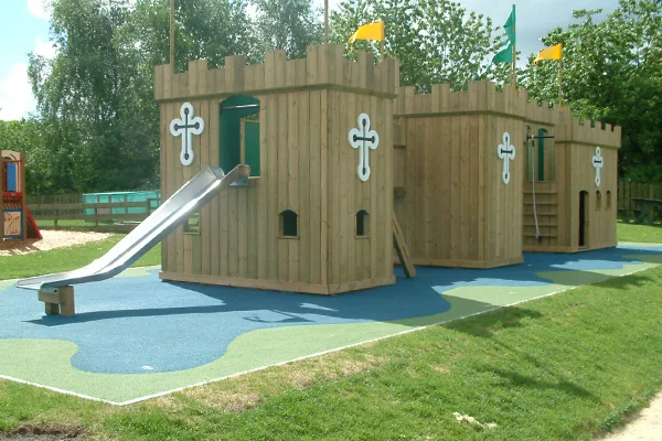 The Dalscone Fort timber play castle featuring a wide stainless steel slide, castle battlements with yellow and green flags, and a white cross emblem, on a blue and green rubber safety surface.