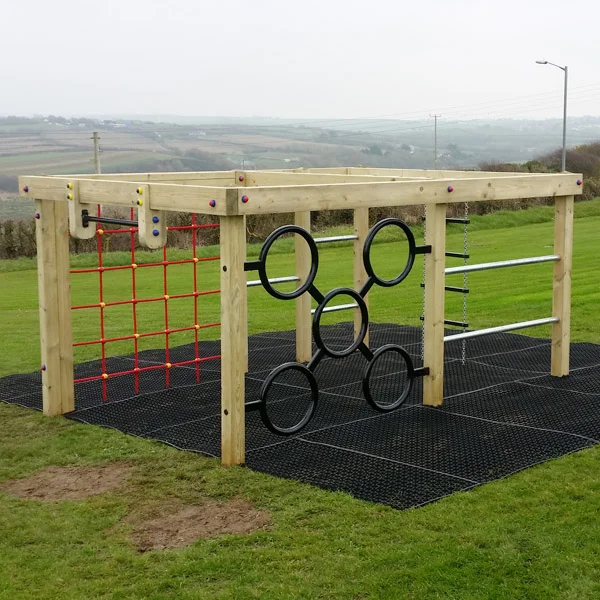 The Delabole Activity Centre timber climbing frame with a green rope net and steel monkey rings, set on black rubber matting in a landscaped outdoor playground with hedges.