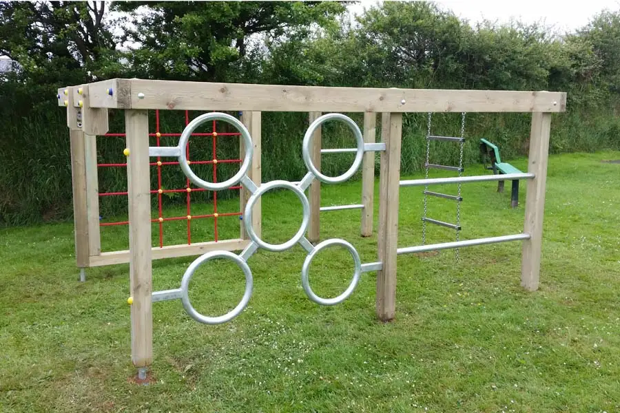 The Delabole Activity Centre timber climbing frame from a different angle, showing the red rope net, a set of steel monkey rings, and a chain ladder, on a grassy surface with trees in the background.