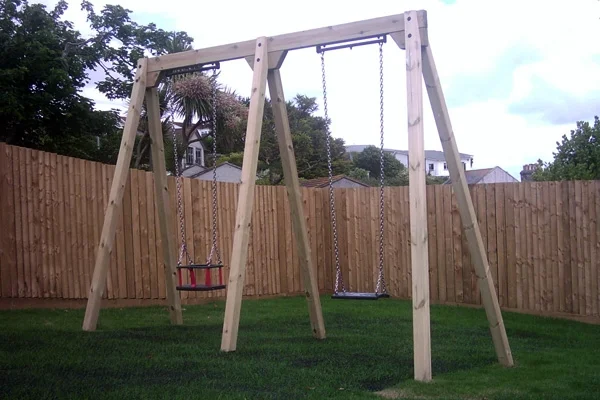 A timber double bay swing with a red toddler seat and a black flat seat, set on a green lawn in a garden with a high wooden fence.