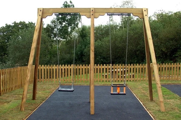 A timber double bay swing with a flat seat and a toddler seat, set on a black rubber path in a woodland playground with a picket fence.