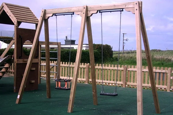A timber double bay swing with a toddler seat and a flat seat, next to a play tower on a green rubber surface in a fenced playground.