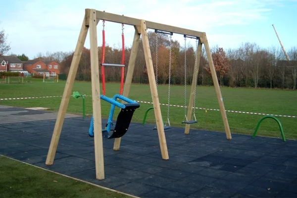 A timber double bay triple swing with a large inclusive seat and one flat seat, on a black rubber tile surface in a park.