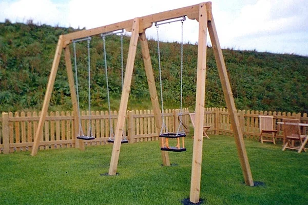 A timber double bay triple swing with two flat seats and one toddler seat, set on a grassy area in a playground with a picket fence.