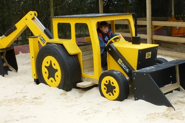 A yellow and black timber play JCB digger with a boy inside, in a sand pit at an outdoor playground.